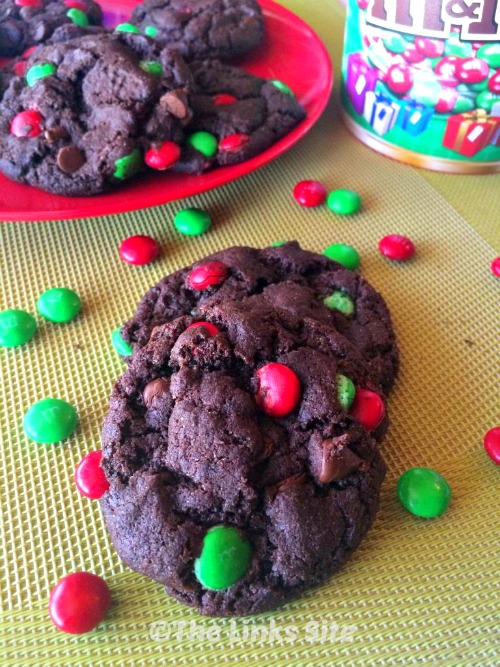 Two cookies on a baking mat with red and green M&M's scattered around them. A red plate with more cookies as well as a bucket of M&M's can be seen in the background.