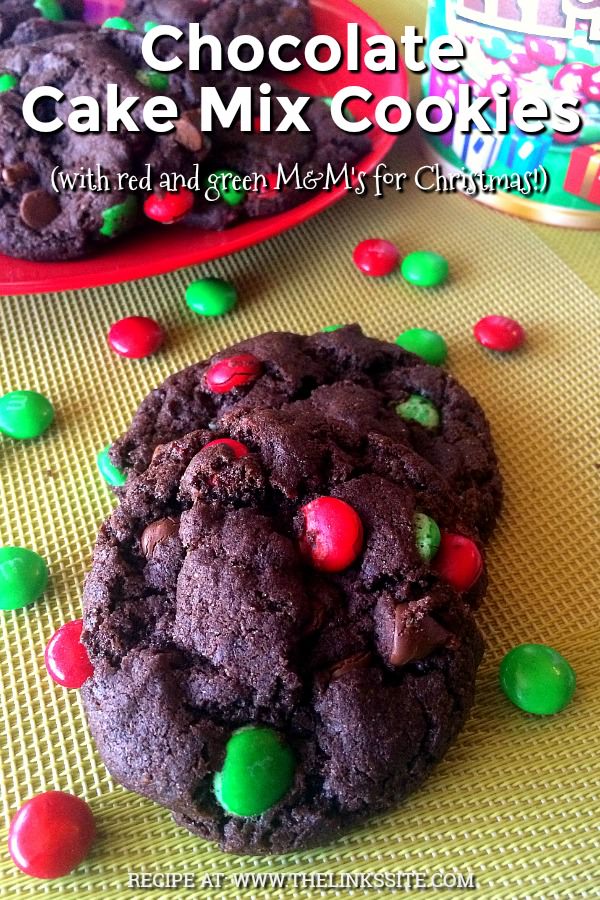 Two cookies on a baking mat with red and green M&M's scattered around them. A red plate with more cookies as well as a bucket of M&M's can be seen in the background. Text overlay says: Chocolate Cake Mix Cookies (with red and green M&M's for Christmas!).