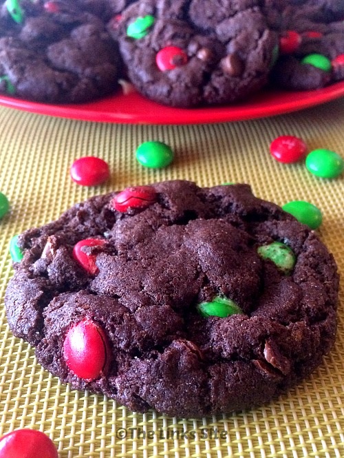 One chocolate M&M cookie on a silicone baking mat with a plate containing more cookies in the background. Several red and green M&M's are scattered around.