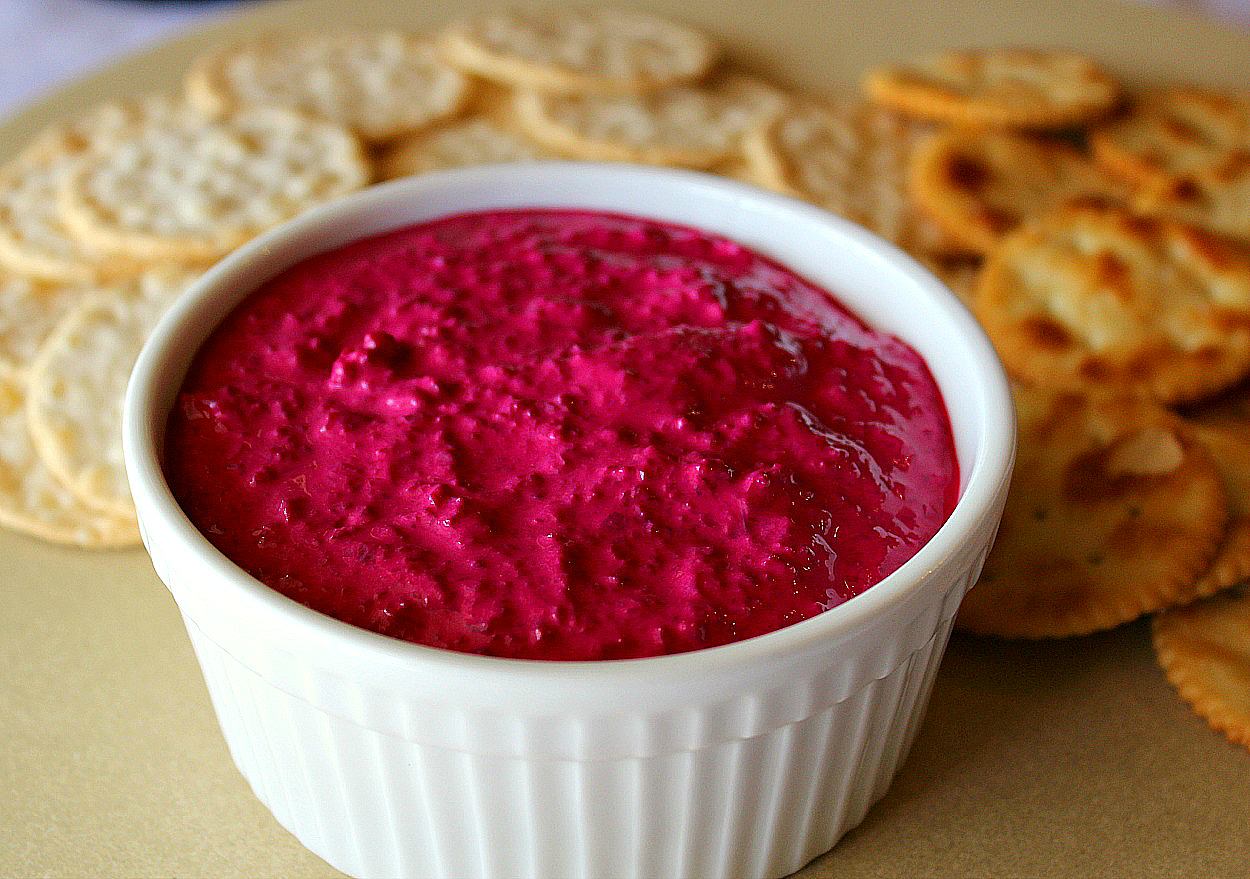 A white ramekin filled with beetroot dip and two types of crackers are arranged on a beige platter.