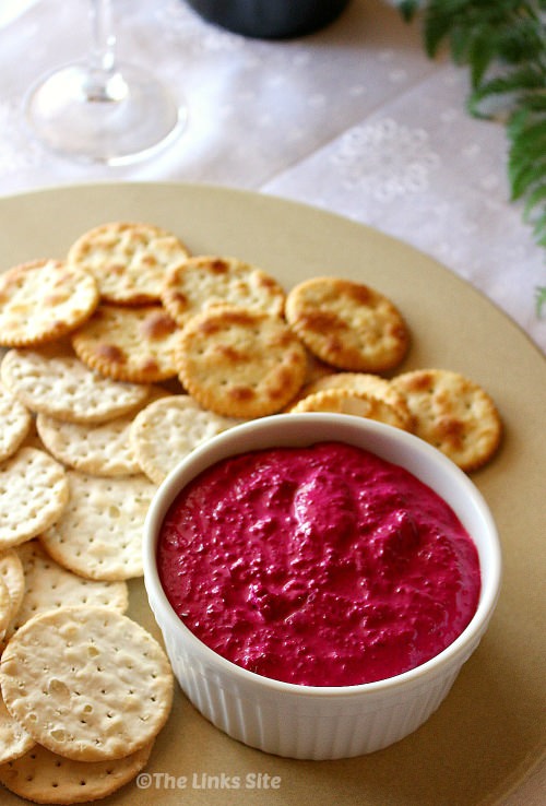 A white ramekin filled with beetroot dip and two types of crackers are arranged on a beige platter. The bottom of a wine glass can be seen in the background.