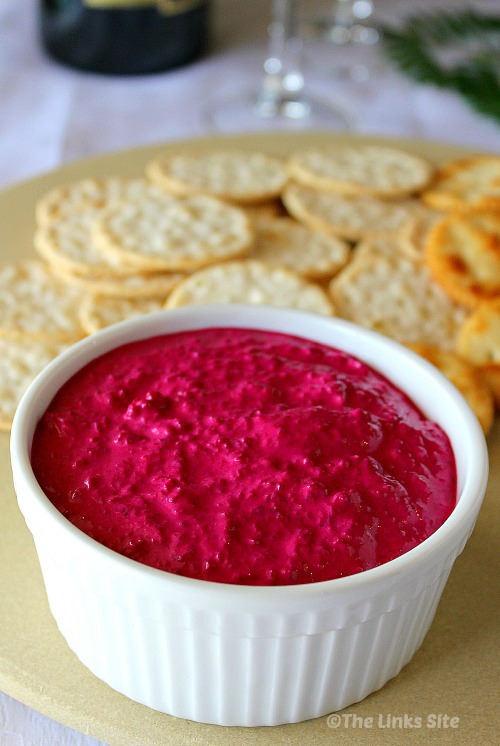 A white ramekin filled with beetroot dip can be seen in the foreground and two types of crackers can be seen in the background arranged on a beige platter.