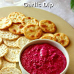 A white ramekin filled with beetroot dip and two types of crackers are arranged on a beige platter. The bottom of a wine glass can be seen in the background and a text overlay reads: 3 Ingredient Roasted Beetroot & Garlic Dip.