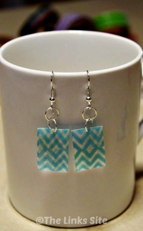 A pair of earrings decorated with blue and white washi tape is pictured hanging from the side of a white coffee mug.