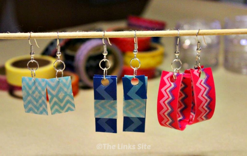 3 Sets of different colored earrings are hanging from a wooden skewer. Rolls of washi tape can be seen in the background.