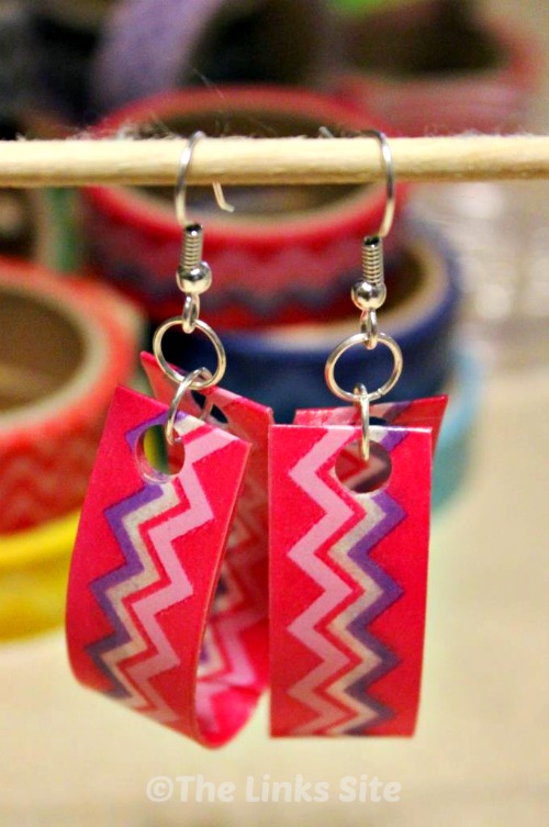 Two earrings decorated with pink, purple, and white zig zag patterned washi tape are pictured hanging from a wooden skewer. 