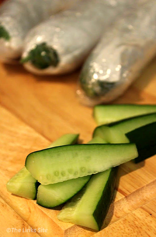 Thick cut strips of cucumber on a wooden chopping board. Whole cucumbers wrapped for storage can be seen in the background.