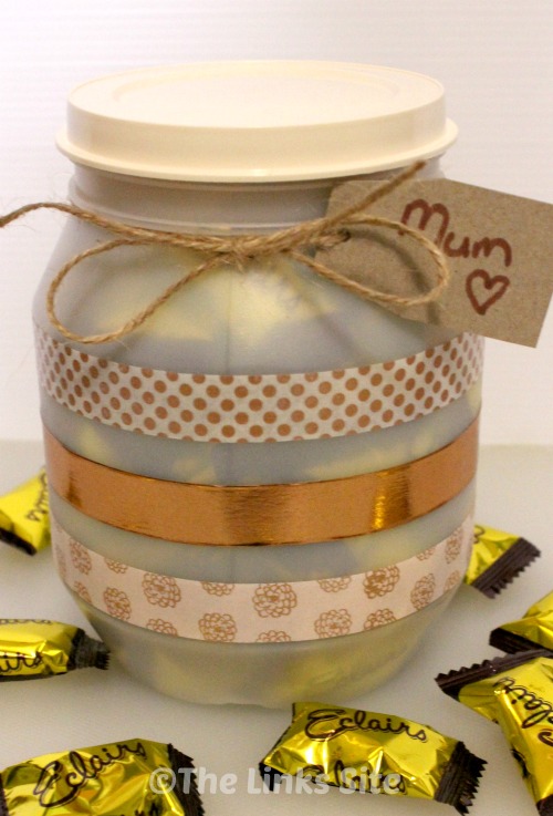 Plastic container with lid that has been decorated with washi tape in gold tones. The container has a gift tag addressed to Mum attached and there are Éclair lollies scattered around the base.
