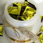 Looking down into the top of a plastic container that is filled with Éclair lollies. The container is decorated with washi tape and extra lollies are scattered around the outside. A gift tag address to Mum is attached with twine.