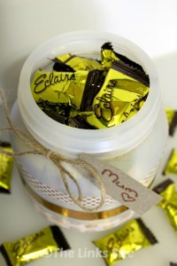 Looking down into the top of a plastic container that is filled with Éclair lollies. The container is decorated with washi tape and extra lollies are scattered around the outside. A gift tag address to Mum is attached with twine.
