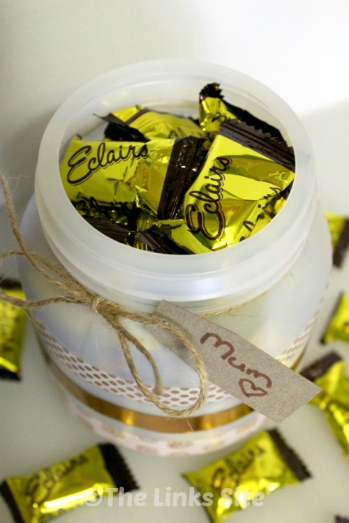 Empty plastic container that has been decorated with white and gold washi tape. The container is filled with and surrounded by sweets and an cardboard gift tag with 'Mum' is attached with twine.