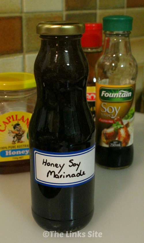 A bottle filled with dark coloured marinade and labelled ‘Honey Soy Marinade’ is placed on a bench. Some of the ingredients used to make the marinade can be seen in the background.
