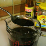 A glass measuring jug is filled with a very dark brown marinade. A large metal spoon has scooped up some of the marinade and is hovering over the jug. Some of the ingredients used to make the marinade can be seen in the background. Text overlay says: Easy Honey Soy Marinade (great with chicken, beef, or lamb!).