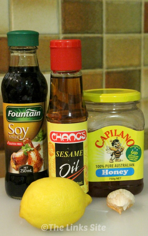 The ingredients used in this recipe are pictured on a bench. Image shows a bottle of soy sauce, a bottle of sesame oil, a jar of honey, a whole lemon, and a clove of garlic.