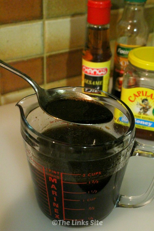 A glass measuring jug is filled with a very dark brown marinade.  A large metal spoon has scooped up some of the marinade and is hovering over the jug. Some of the ingredients used to make the marinade can be seen in the background.
