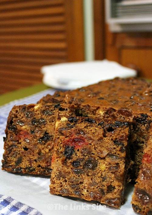 Fruit cake, with some pieces cut, on a table with white plates and brown cupboards in the background. 