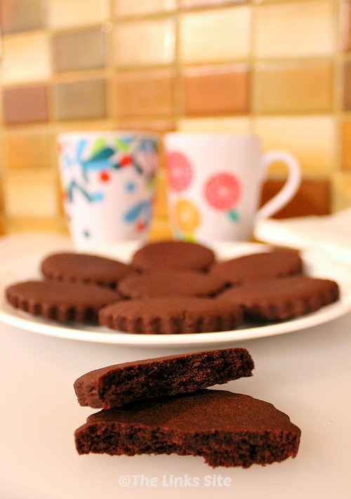 One broken chocolate sugar cookie is seen in the foreground and a plate containing more cookies plus two mugs are in the background.