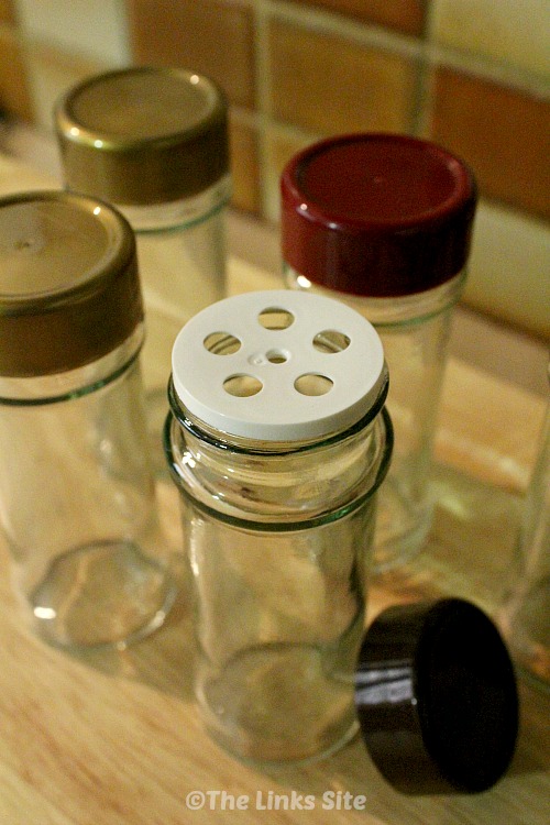 Four empty glass spice jars are pictured on a wooden cutting board. One of the jars has its plastic lid removed to show the white plastic shaker insert.