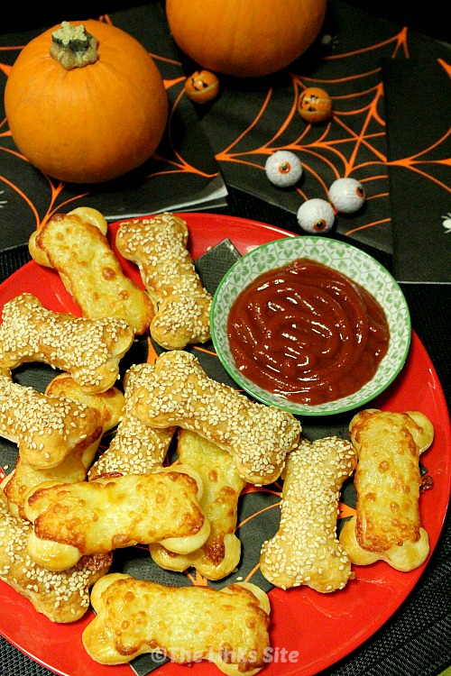 Overhead image of a plate of pastry bones and a small bowl of tomato sauce. Halloween decorations including two small pumpkins can be seen in the background.