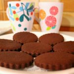 White plate containing several scalloped edged chocolate sugar cookies. There are two mugs in the background.