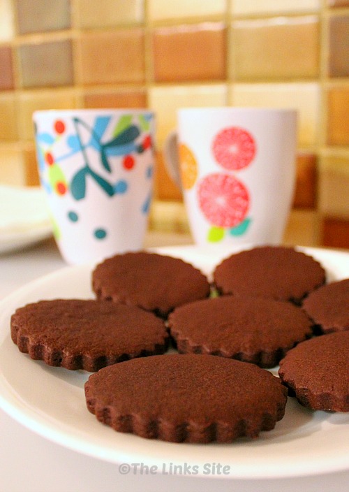 White plate containing several round, scalloped edged chocolate sugar cookies. There are two mugs in the background.
