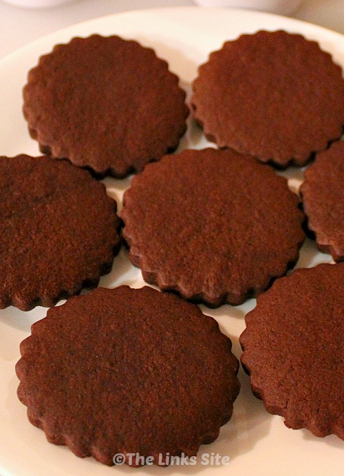 Close up of seven scalloped edged chocolate sugar cookies on a white plate.