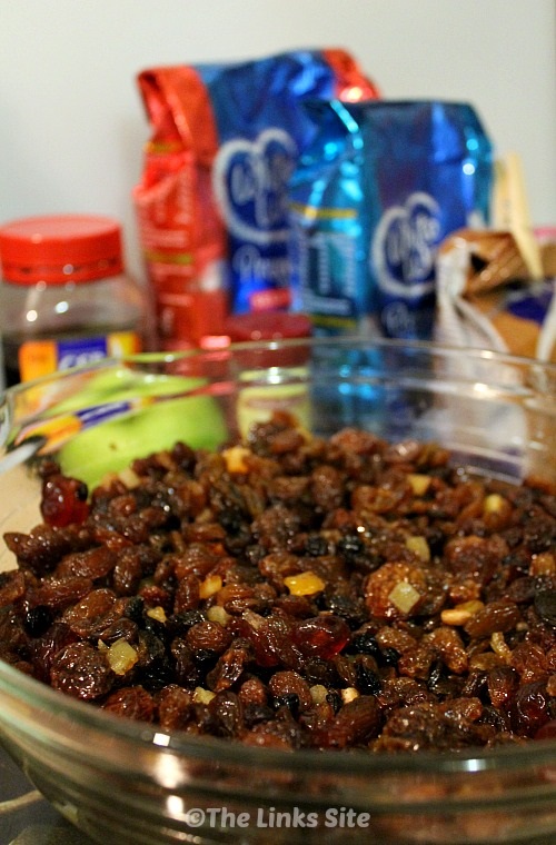 Glass bowl filled with mixed dried fruit. Other cake ingredients such as flour, sugar, and spices can be seen in the background.