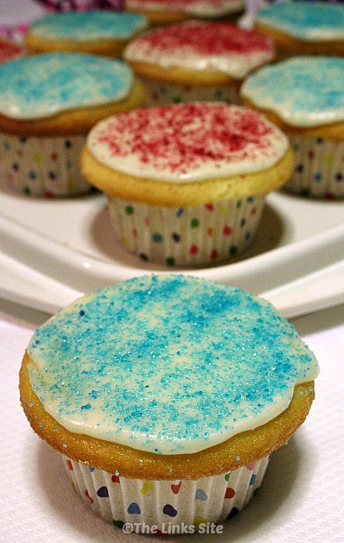 One cupcake decorated with white icing and blue sugar glitter is in the foreground. More cupcakes decorated with pink or blue sugar glitter can be seen in the background.