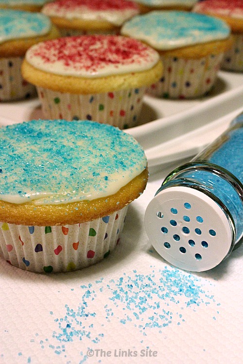 One cupcake decorated with white icing and blue sugar glitter is in the foreground. Alongside the cupcake is a spice shaker filled with blue sugar glitter that has been knocked over and is spilling some sugar out. More decorated cupcakes can be seen in the background.