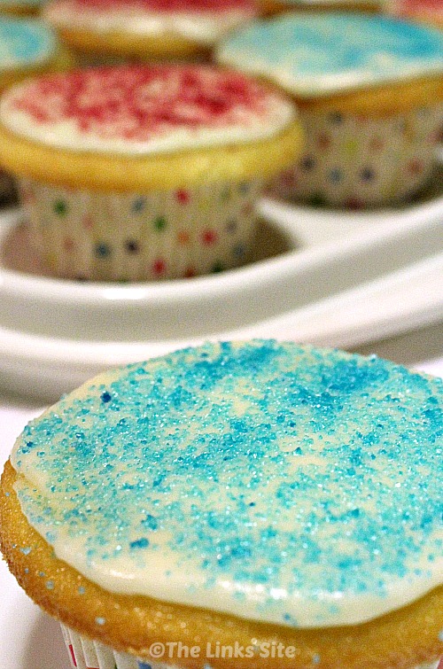 Close up of a vanilla cupcake decorated with white icing and blue sugar glitter. More cupcakes can be seen in the background.