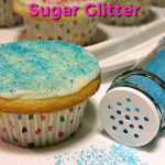 One cupcake decorated with white icing and blue sugar glitter is in the foreground. Alongside the cupcake is a spice shaker filled with blue sugar glitter that has been knocked over and is spilling some sugar out. More decorated cupcakes can be seen in the background. Text overlay says: How to Make Your Own Sugar Glitter.