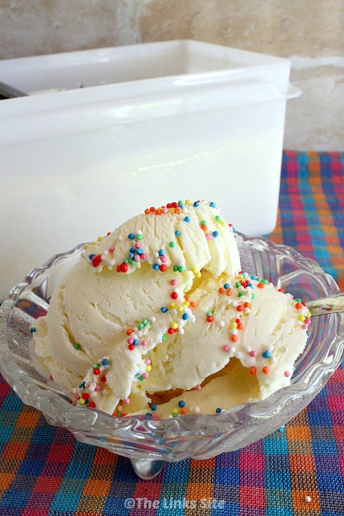 Vanilla ice cream topped with multi coloured sprinkles in a glass bowl. The bowl is placed on a pink, blue, and orange check cloth with a plastic ice cream tub in the background.
