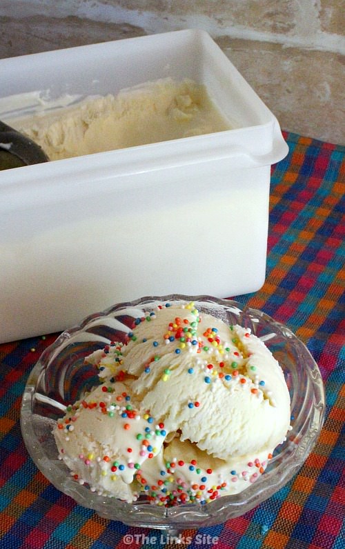 Overhead view of some vanilla ice cream topped with multi coloured sprinkles in a glass bowl. The bowl is placed on a pink, blue, and orange check cloth with a plastic ice cream tub in the background.