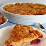 A piece of torte on a white plate is pictured in the foreground. The rest of the torte in a white baking dish, along with a silver spoon and a small bowl of plums, can be seen in the background.