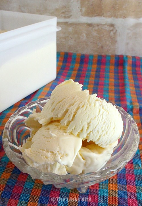 3 Scoops of vanilla ice cream in a glass bowl. The bowl is placed on a pink, blue, and orange check cloth with a plastic ice cream tub in the background.