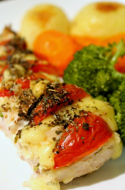 A cooked piece of chicken that is topped with generous amounts of tomato, herbs, and cheese and placed on a white plate. Vegetables can be also seen on the plate behind the chicken.