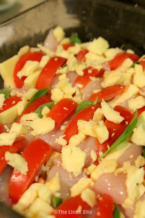 Raw chicken fillets in a glass baking dish. The tops of the fillets have been stuffed with tomato, basil, and cheese. Extra cheese is also sprinkled on top.
