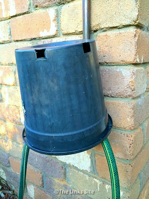 A black plastic plant pot has been placed over a garden tap. Two pieces of hose can be seen coming out from under the pot. The tap is located against a brick wall.
