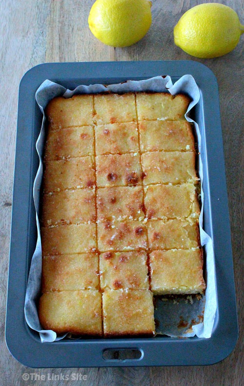 Overhead view of a lemon cake in a lined baking pan. The cake has been cut into squares and one corner piece is missing. The baking pan is resting on a wooden board and there are two lemons next to the baking pan.