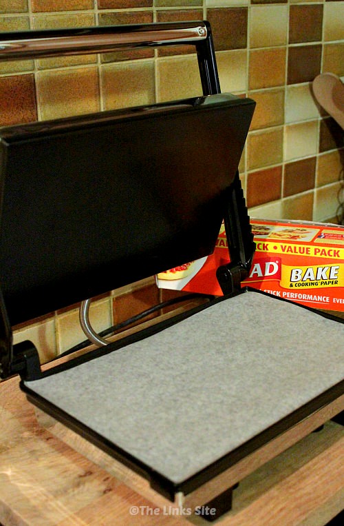 Open sandwich press with baking paper lining the bottom hotplate. A boxed roll of baking paper can be seen in the background.
