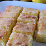 Squares of lemon cake on a white plate. Two lemons can be seen in the background. Text overlay says Easy Lemon Cake (with crunchy lemon drizzle topping!).