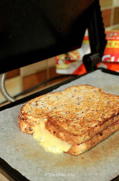 Toasted sandwich sitting on the plate of a sandwich press. Melted cheese is oozing out onto the baking paper lined hotplate.
