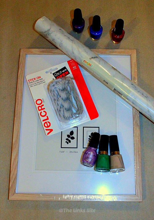 Overhead view of a photo frame, a packet of Velcro dots, a roll of adhesive vinyl, and six bottles of nail polish on a work bench.