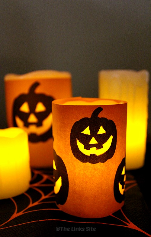 Four battery powered pillar candles are pictured on black surface that is decorated with a spider web pattern. Two of the candles have been wrapped with orange craft paper that is decorated with black Jack O Lantern faces so that the light from the candles shines through the facial features. 
