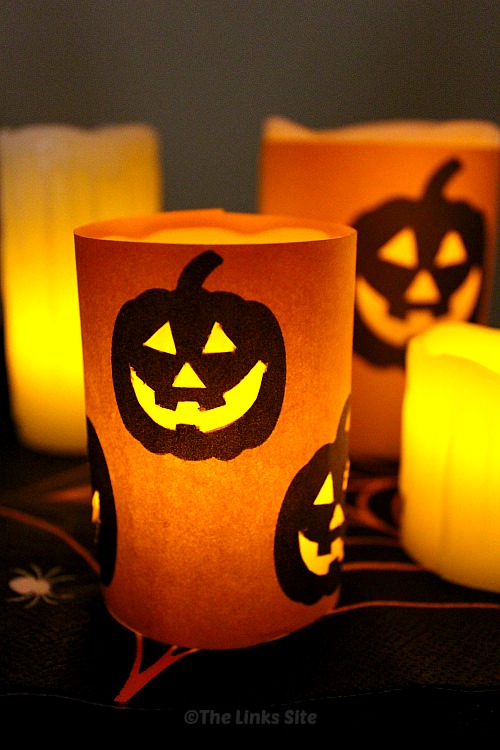 Dimly lit image showing battery powered pillar candles wrapped with orange craft paper that is decorated with Jack O Lantern faces. The facial features have been cut out so that the light from the candles shines through.