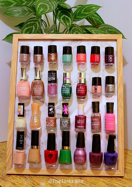Photo frame with light wooden edges sitting on a bench with a potted plant in the background. Four rows of seven nail polish bottles are stuck to the photo frame.