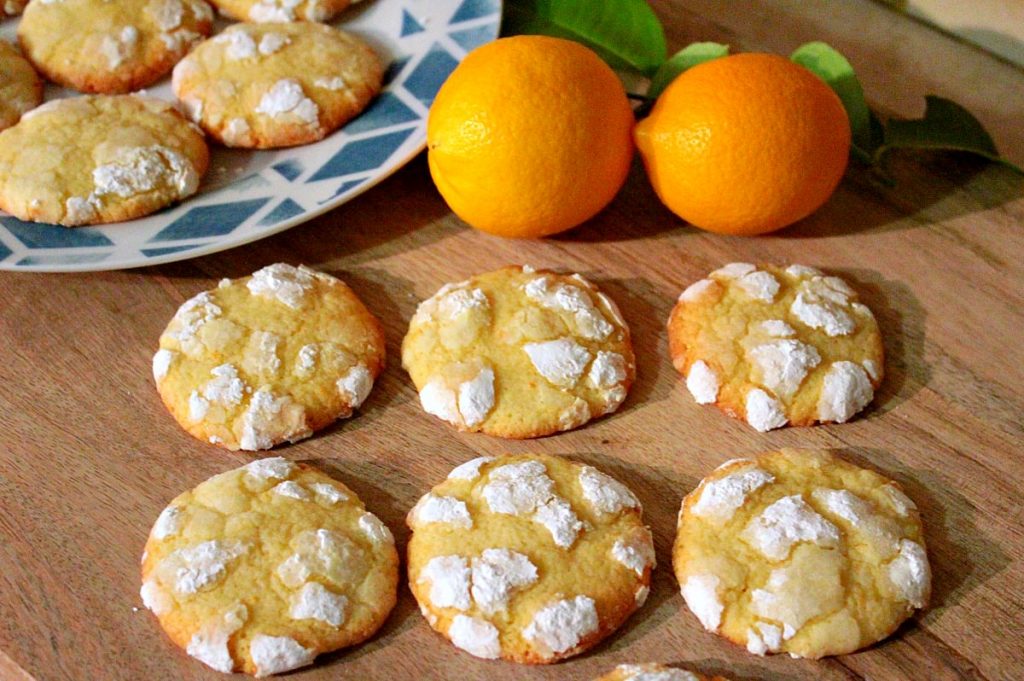 Six crinkle cookies arranged on a wooden board. There is a plate with extra cookies plus two lemons in the background.