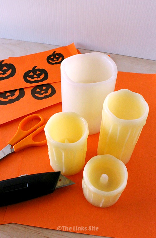 Image shows the materials needed for this project. Pictured are four battery powered pillar candles, orange craft paper, scissors, a utility knife, and Jack O Lantern faces printed out onto some craft paper.