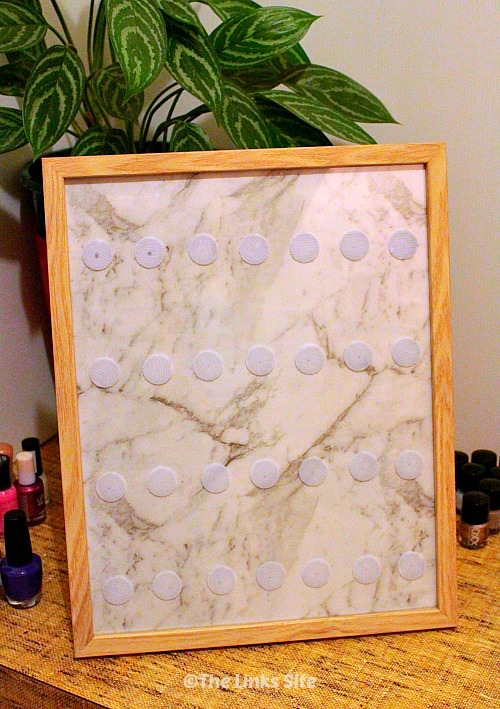 Photo frame with light wooden edge sitting on a bench with nail polish bottles and a potted plant in the background. The photo frame has four rows of seven Velcro dots spaced across its surface.
