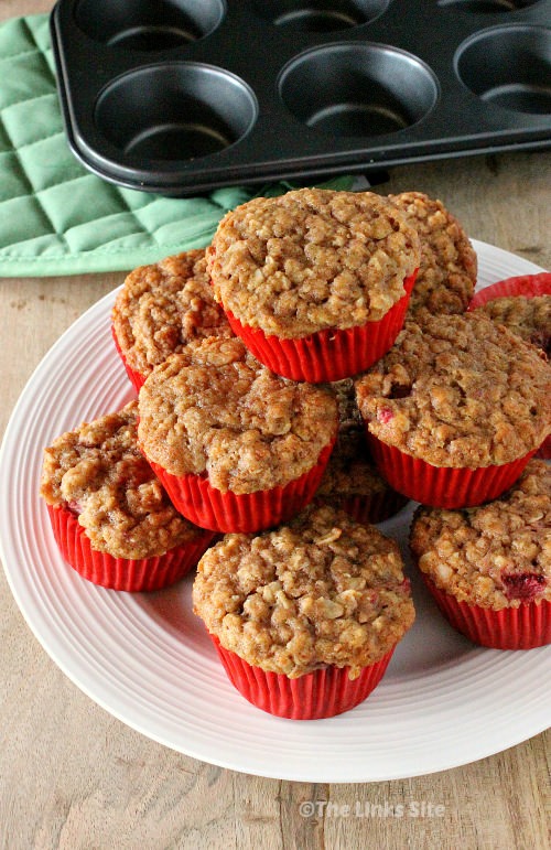 Several muffins in red paper cases are stacked up on a white plate. An empty muffin tin and oven mitt can be seen in the background.